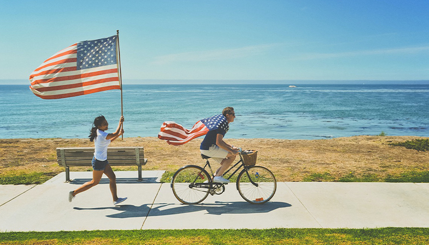 Person running with American flag behind person riding bike with American flag as cape